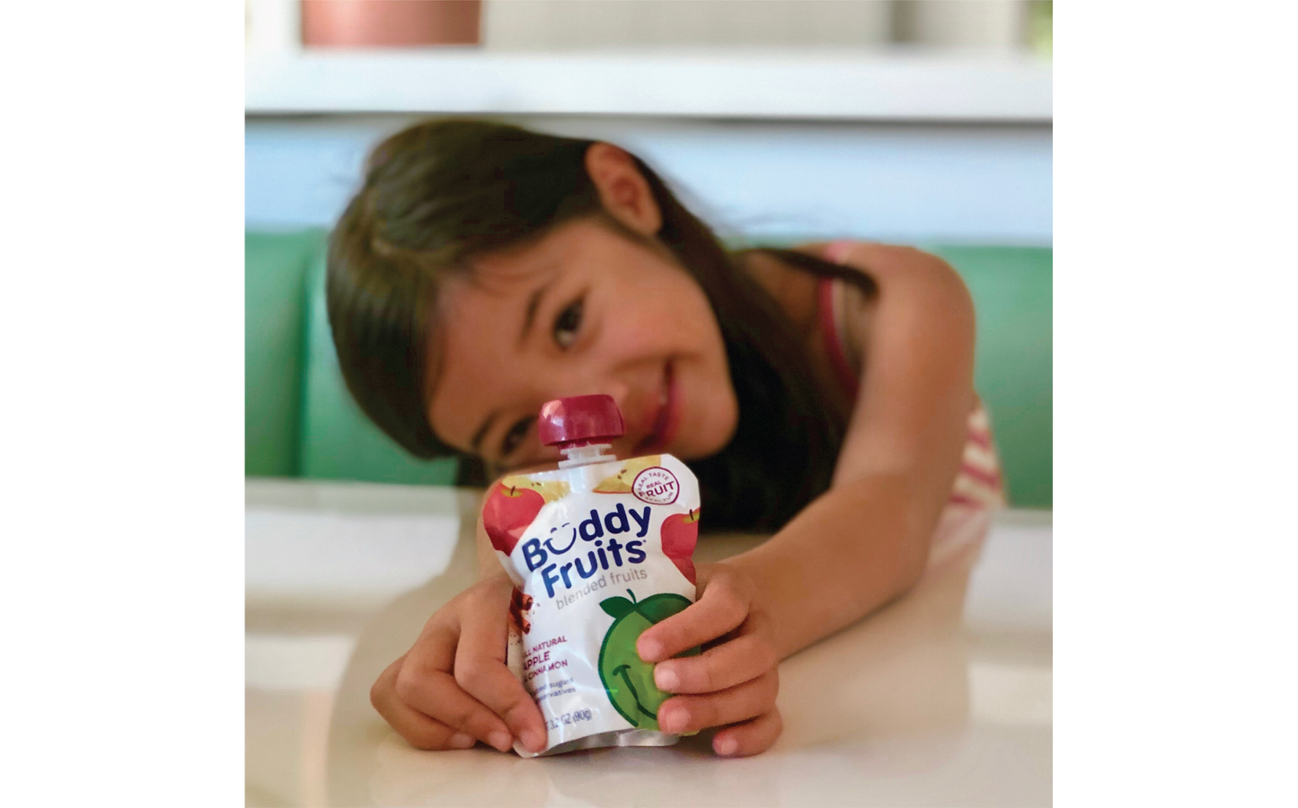 Happy young girl poses with her delicious Buddy Fruits Cinnamon & Apple fruit pouch at the dinner table.
